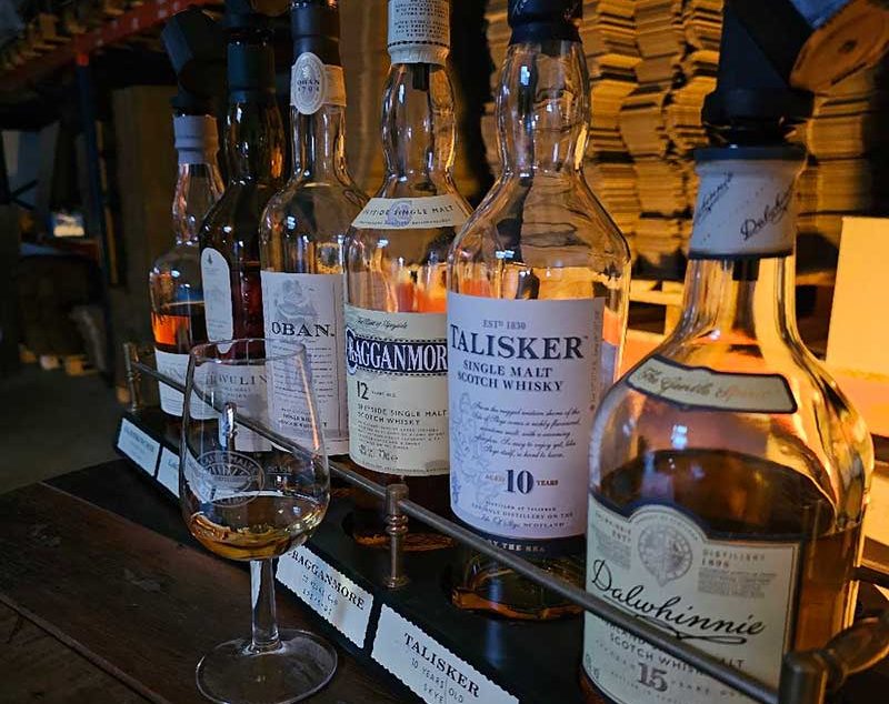 Reihe von Whiskyflaschen mit einem gefüllten Whisky Glas davor in einem dimmbar beleuchteten Raum.