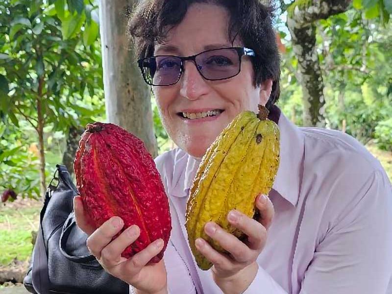 Frau mit Brille hält jeweils eine rote und eine gelbe Kakaoschote in je einer Hand und lächelt.