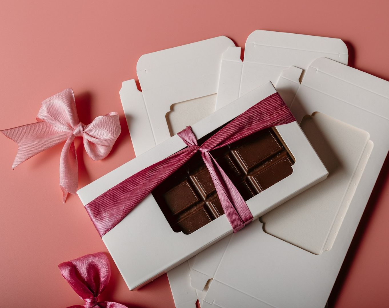 Schokoladenstück in einer weißen Verpackung mit rosa Schleife auf rosa Hintergrund.