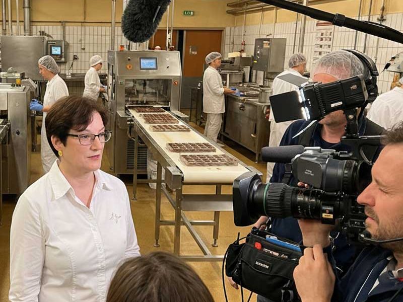 Bildschirm mit zwei Frauen auf der Bühne, eine liest von einem Blatt und spricht in ein Mikrofon.