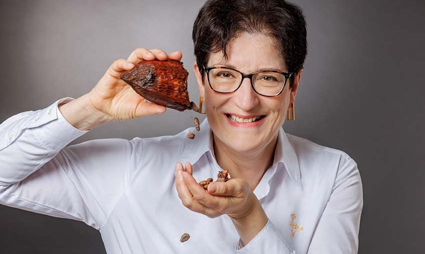 Lächelnde Frau in weißem Hemd hält eine Schokoladenschote und lässt Kakaobohnen heraus fallen.