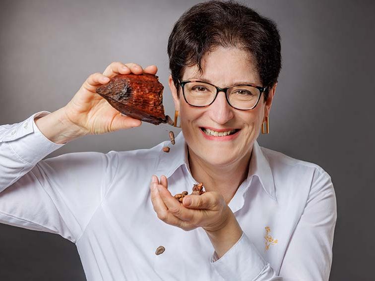 Frau mit Brille lächelt und hält eine Kakaoschote, aus der Kakaobohnen in die andere Hand herausfallen.