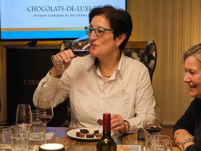 Frau genießt Wein und Schokolade, umgeben von Gästen an einem Tisch.