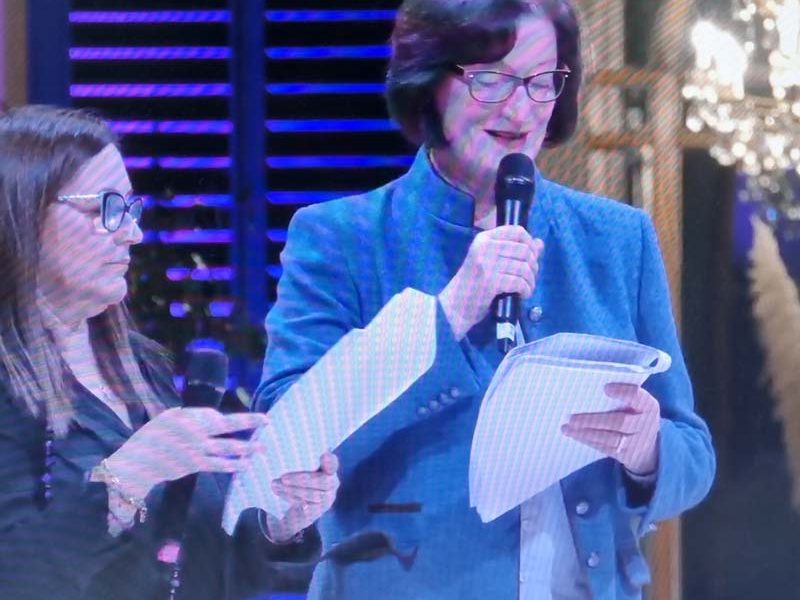 Bildschirm mit zwei Frauen auf der Bühne, eine liest von einem Blatt und spricht in ein Mikrofon.