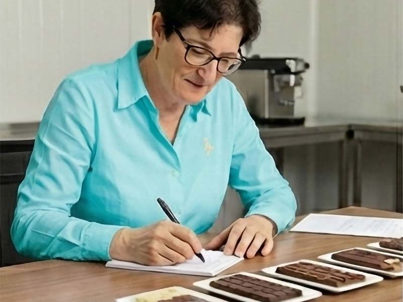 Frau in türkisfarbener Bluse sitzt am Schreibtisch und macht  Notizen zu Schokoladentafeln 