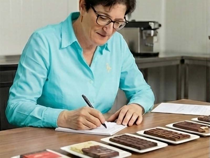 Frau in türkisfarbener Bluse sitzt am Schreibtisch und macht  Notizen zu Schokoladentafeln 