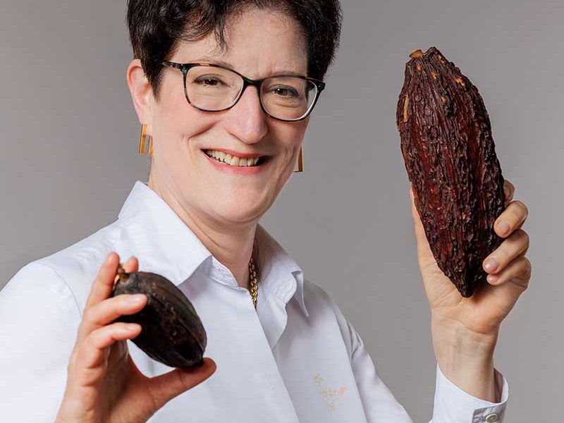 Eine Frau hält eine Kakaoschote in einer Hand und eine kleinere Kakaobohne in der anderen.